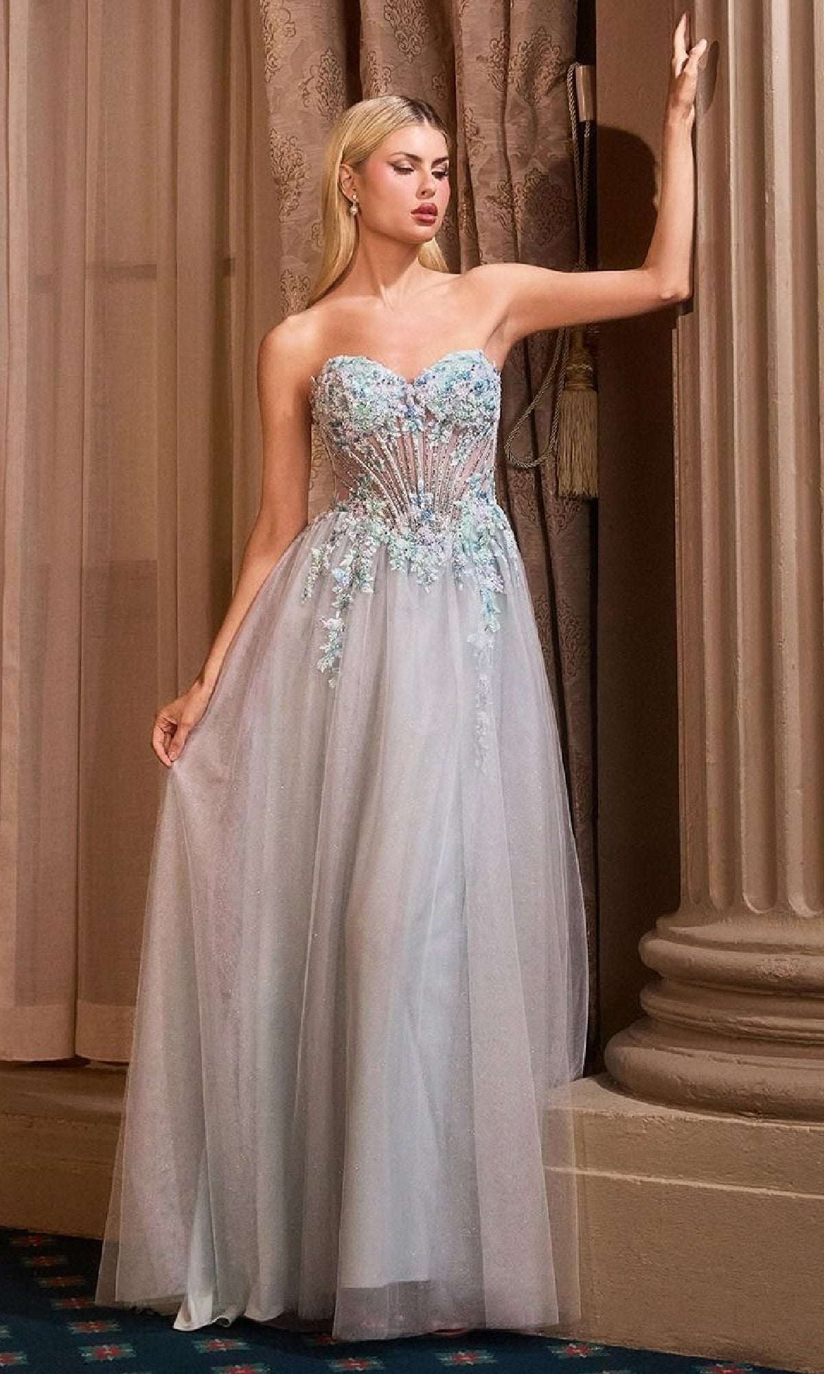 A woman stands indoors in a formal strapless blue beaded prom gown—Andrea & Leo A1478—featuring a sheer skirt and floral-embellished bodice.